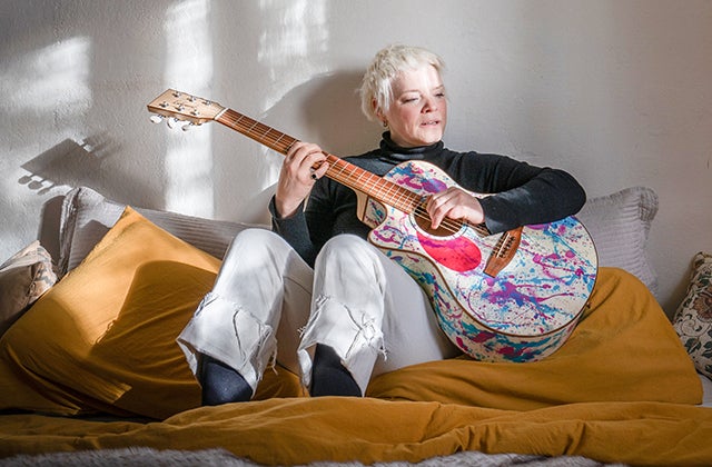 A woman seated on a bed playing a painted splattered guitar.