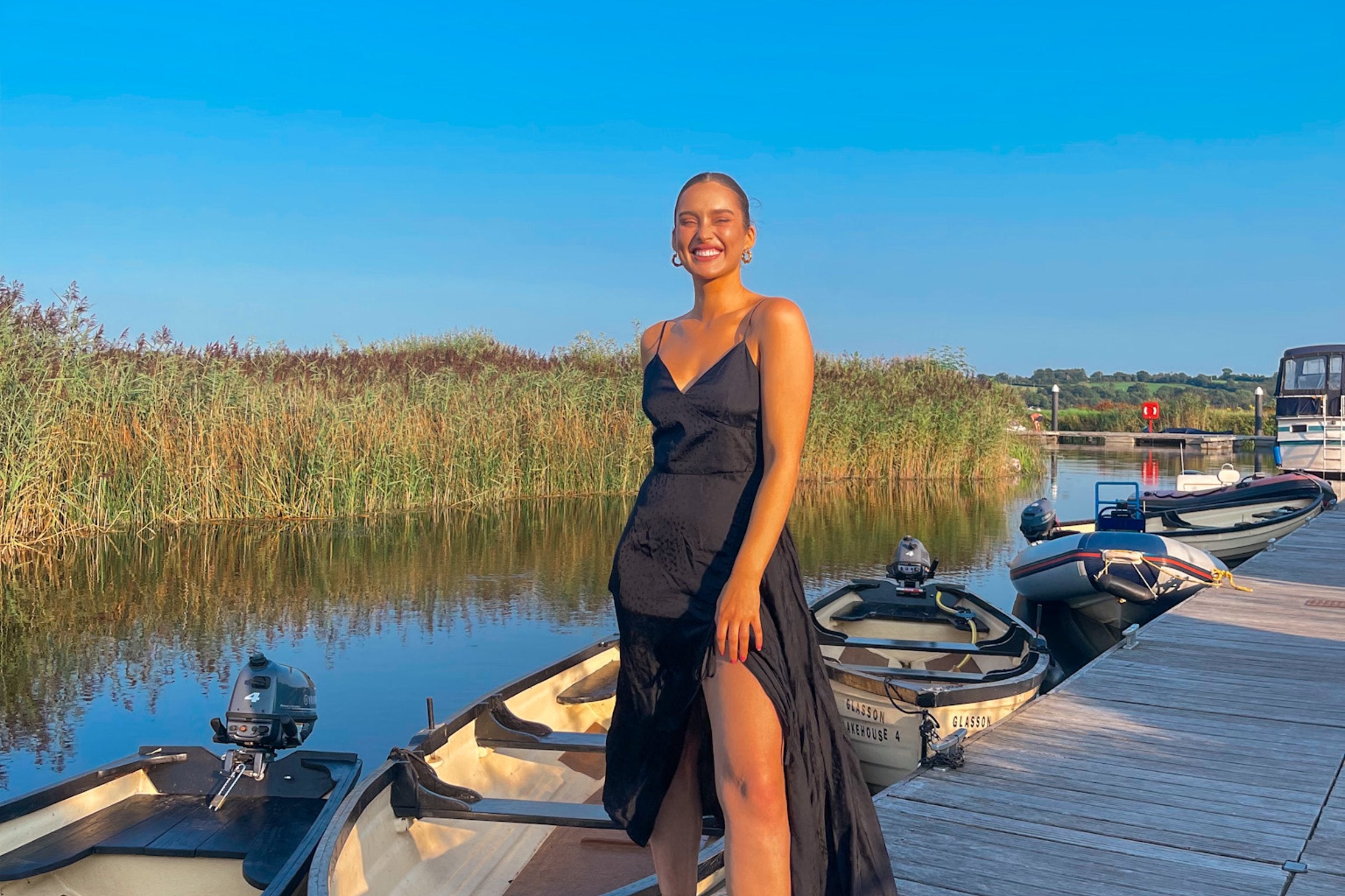 Roz Purcell wearing a black dress standing on a deck in front of a row of boats at Glasson Lakehouse, Westmeath