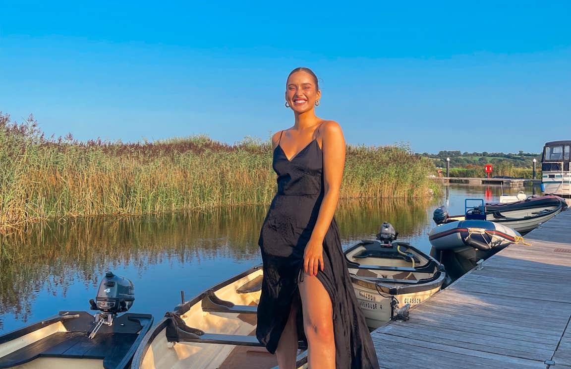 Roz Purcell wearing a black dress standing on a deck in front of a row of boats at Glasson Lakehouse, Westmeath