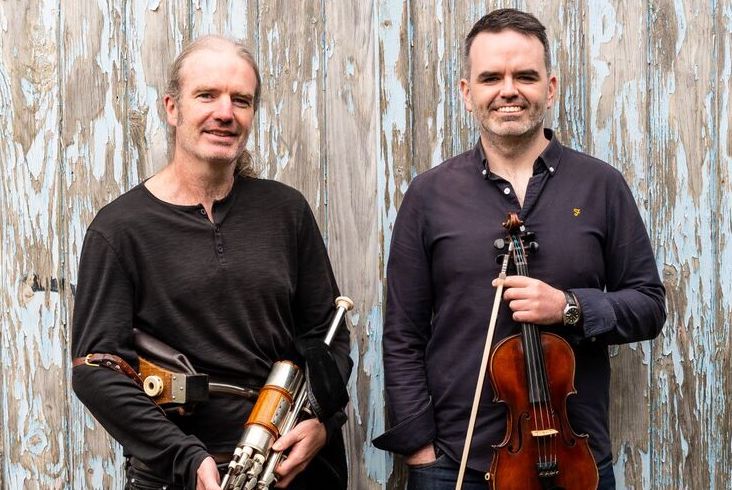 2 men standing beside each other holding musical instruments and smiling, against old weathered fence.