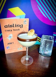 A creamy cocktail in a coupe glass beside an aisling copy book and cassette tapes