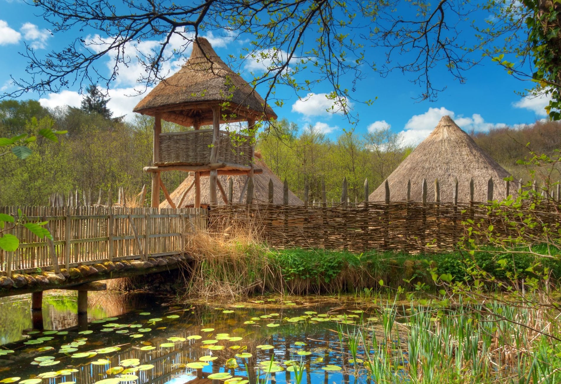A wooden bridge over a lake with a thatched watch tower
