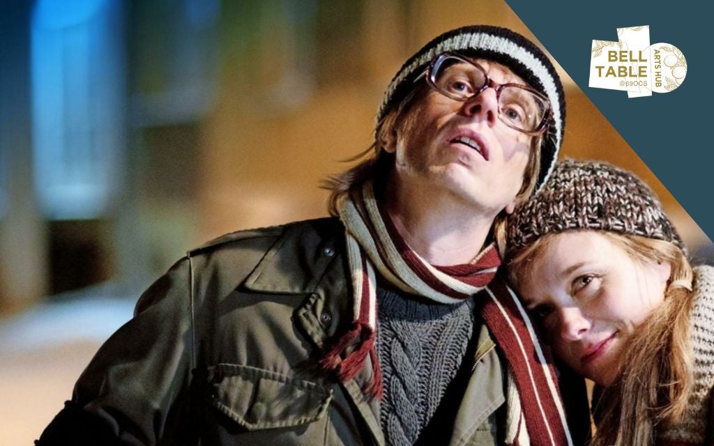 A man in hat and scarf with woman resting her head on his left shoulder.