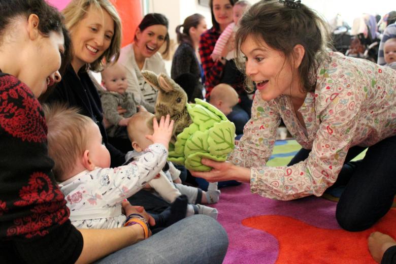 A woman is smiling and holding up a soft toy of a rabbit in a lettuce to a small baby on a parent's lap with others looking on.