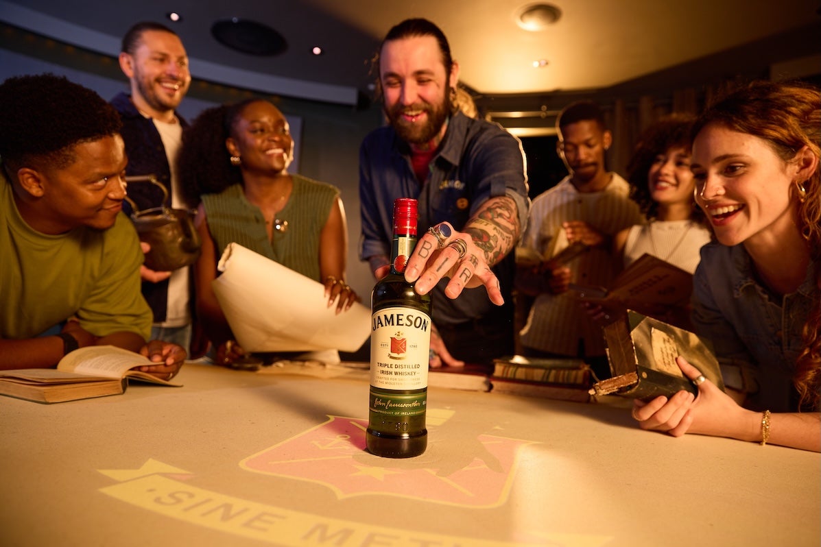 A group of laughing people around a table looking at a bottle of whiskey