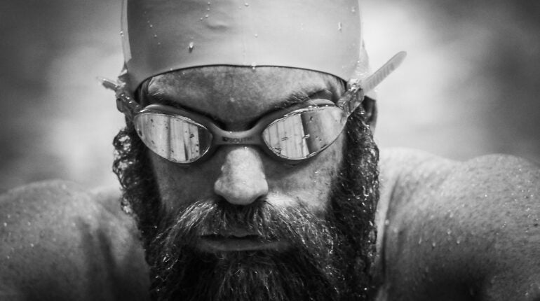 Black and white headshot of man with a beard wearing swim goggles and hat covered in water droplets.