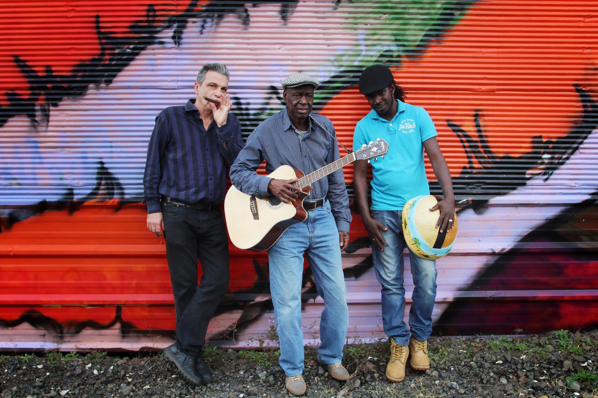 3 men with a guitar and mouthorgan are leaning against a wall painted with red and purple graffiti.