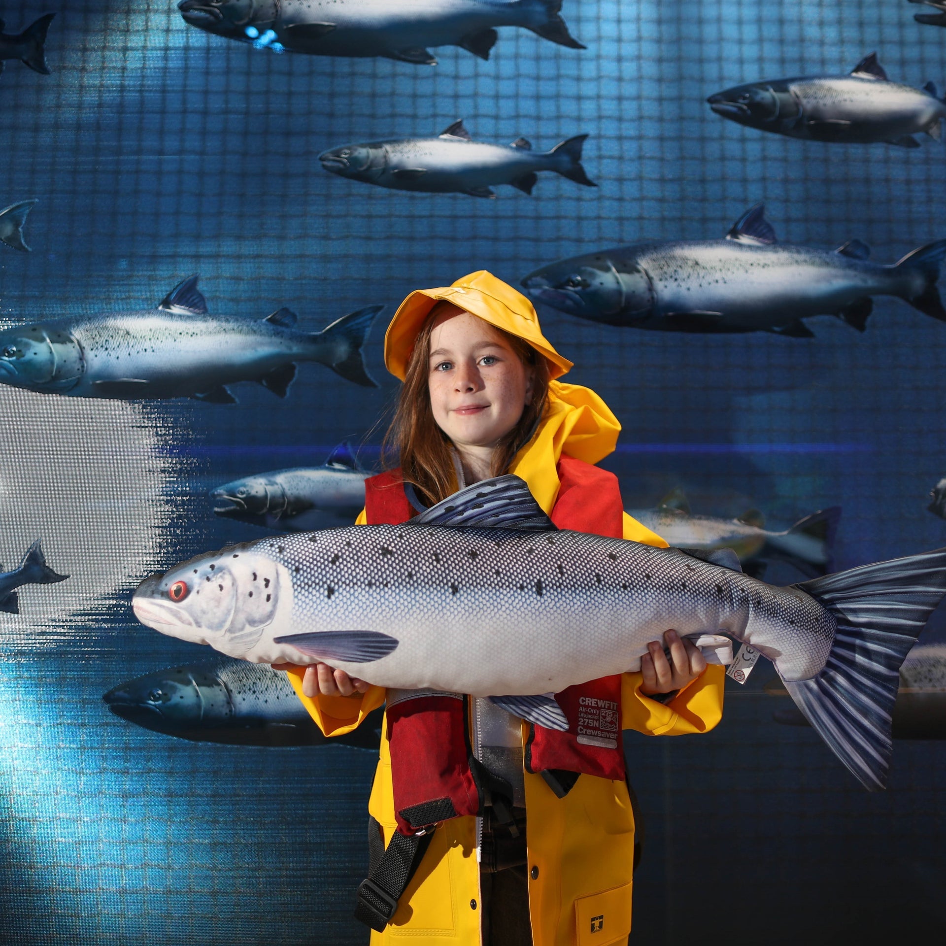 Girl in yellow oil skins holding a toy salmon teddy with salmon swimming in the background