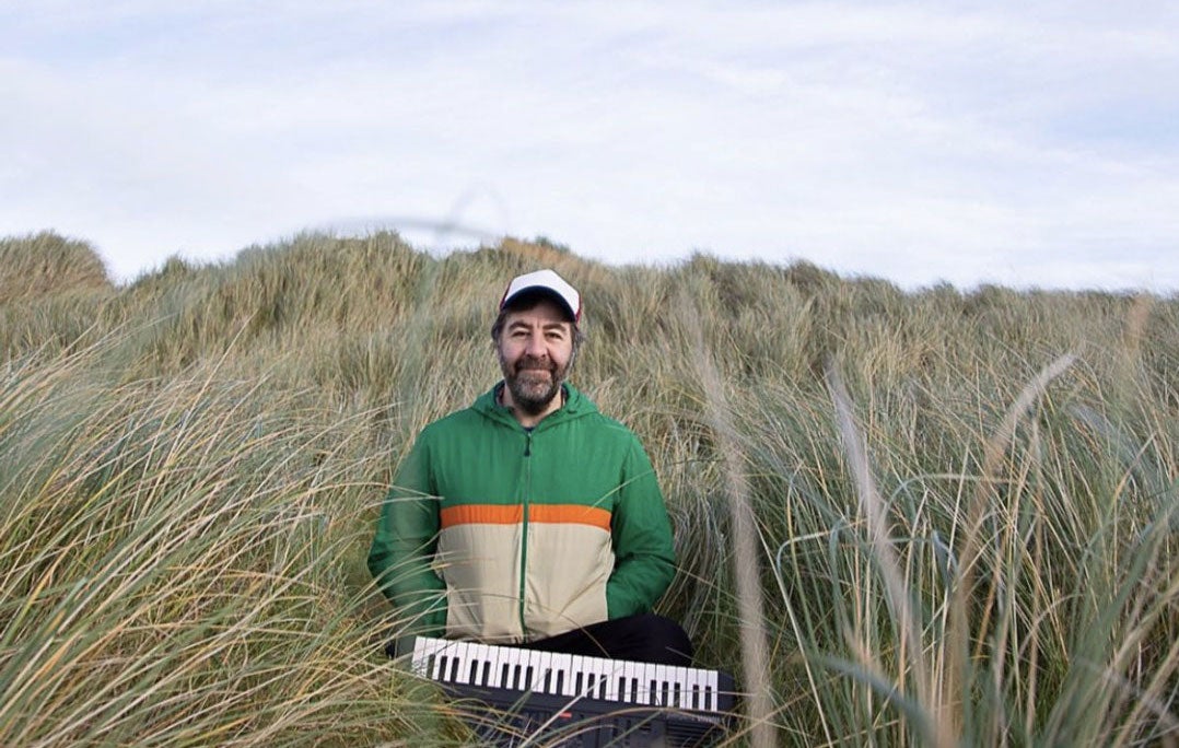 A smiling man is standing in tall grass on a sand dune with a small keyboard in front of him