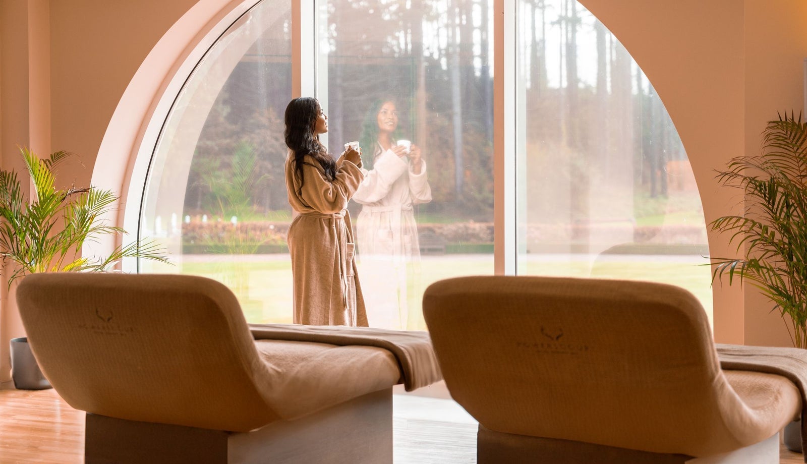 A lady in a bathrobe staring out a large window in a room with treatment beds