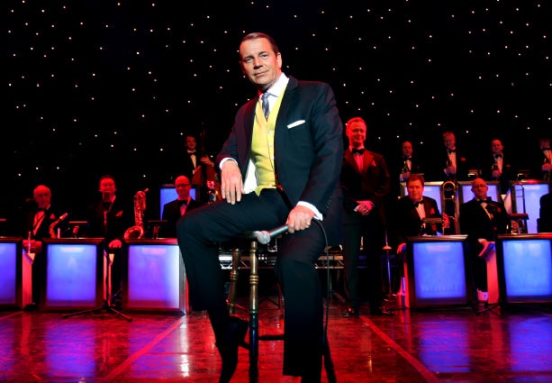 Sinatra's Greatest Hits with Stephen Triffitt. A man in black suit and yellow waistcoat is seated on a stool holding a mic on a stage with band behind him.