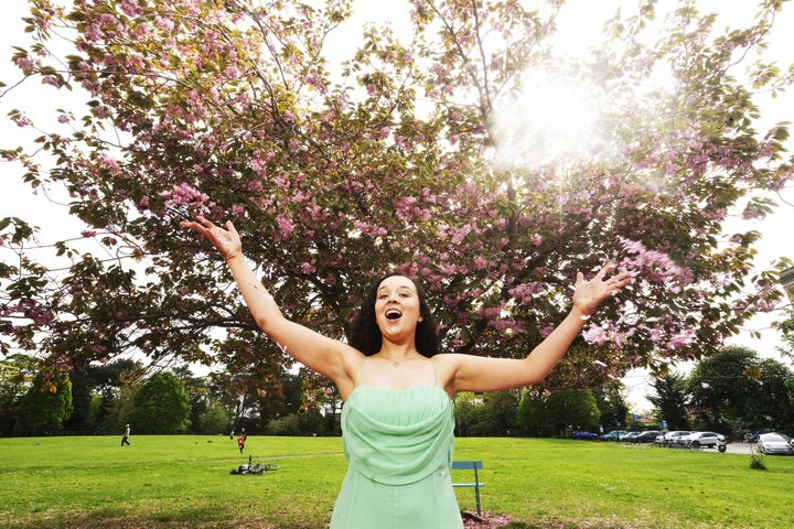 A woman in a green dress with arms outstretched, singing with trees in the background in blossom.