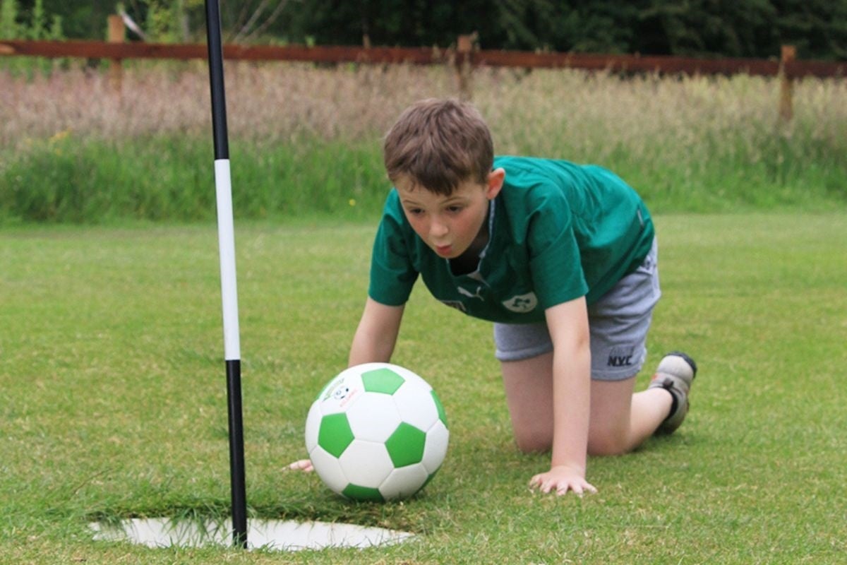 A child on their hands and knees blowing on a football to get it in a hole