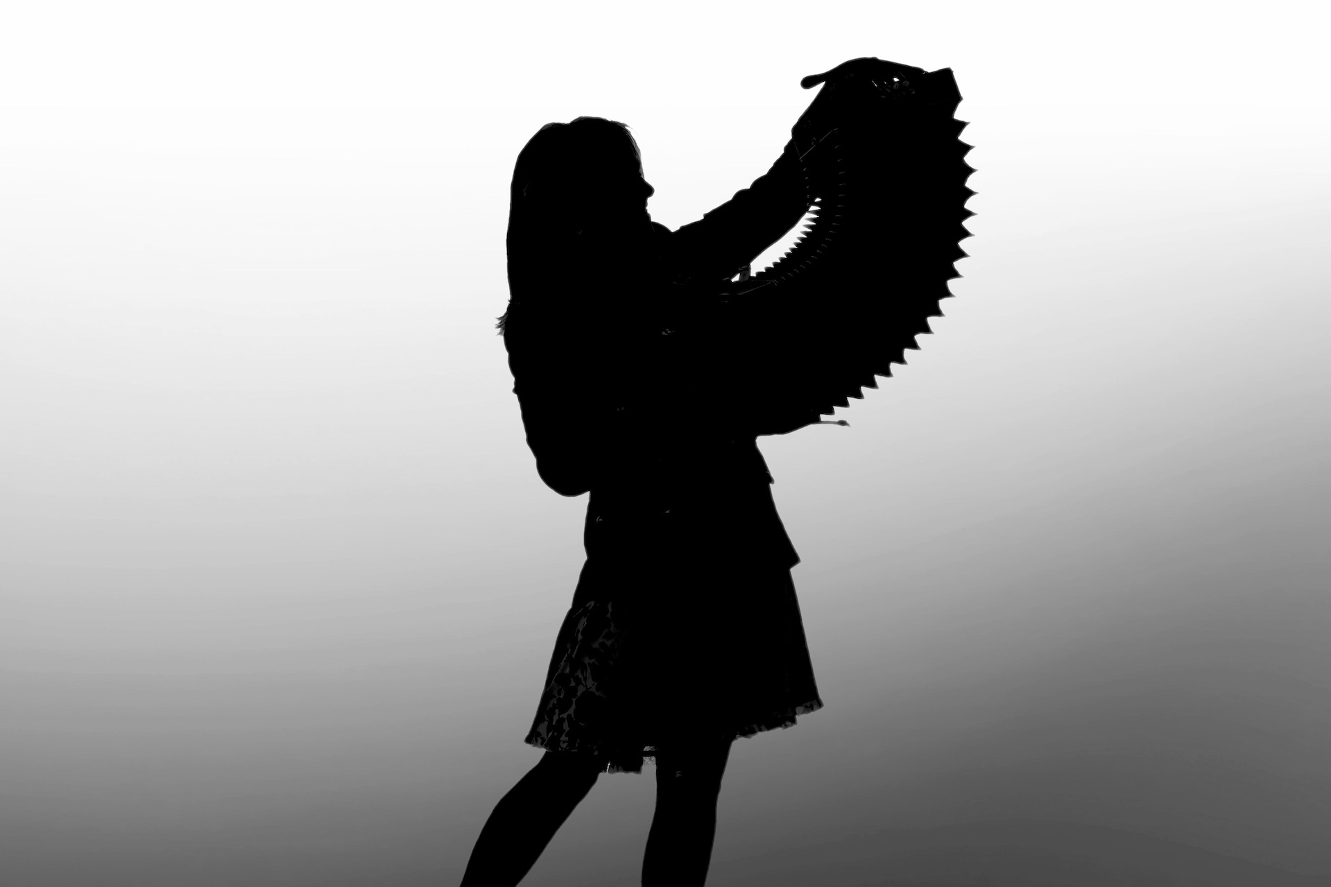 Black silhouette photo of woman in skirt holding up an open accordion against grey background.
