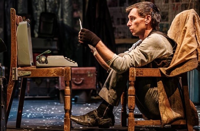 A man seated in a chair frame looking at a piece of paper with old small typewriter on an old chair in front of him.