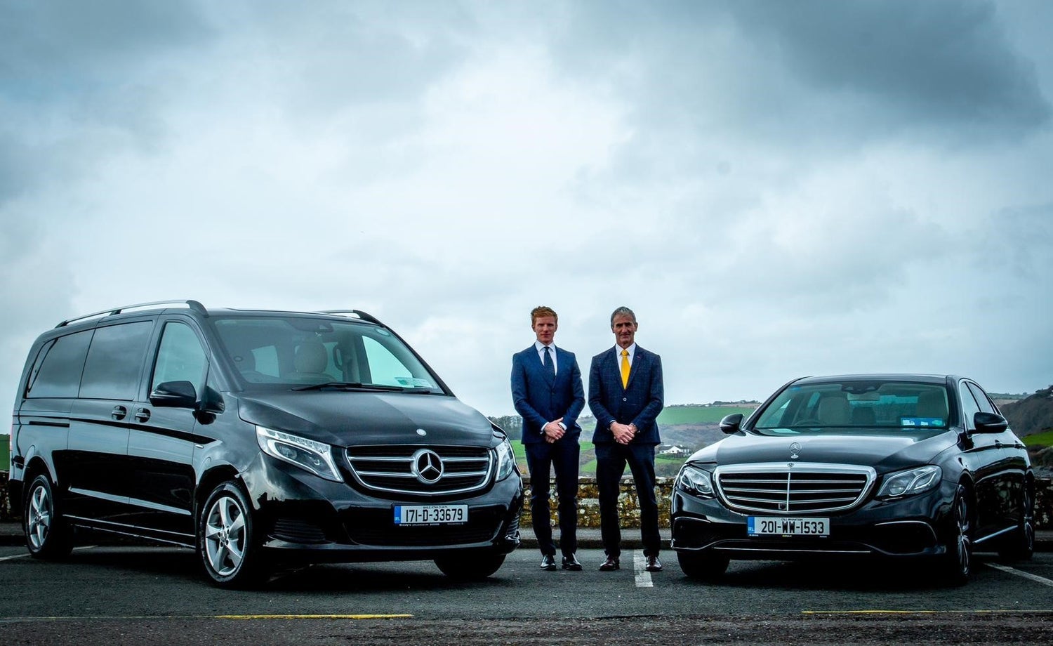 Two people in suits standing between two luxury cars