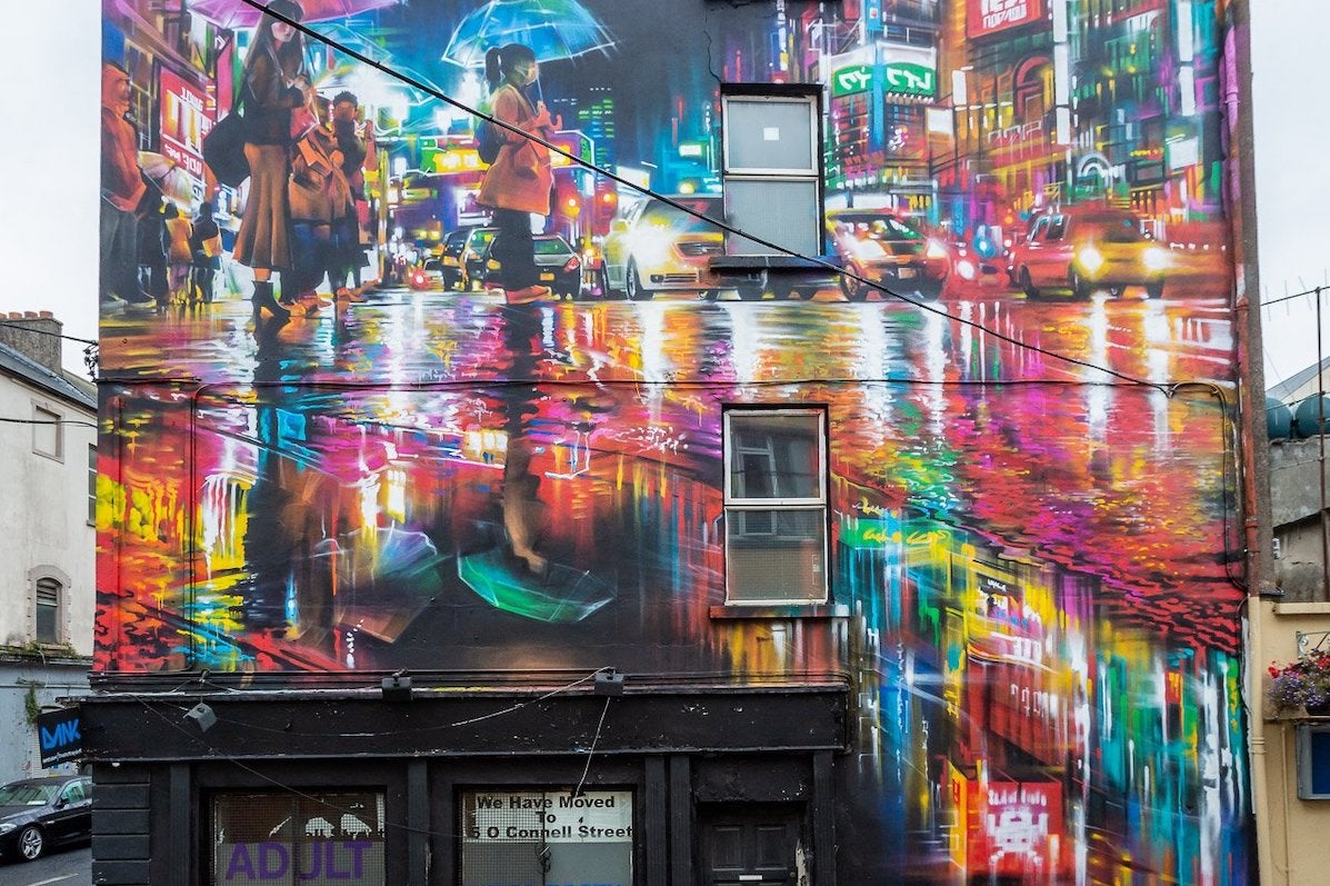 Waterford Walls 2018 - Dan Kitchner. Photographer - Karl Kachmarksy, side of a building painted with a brightly lit city scene at night with reflections in wet road.