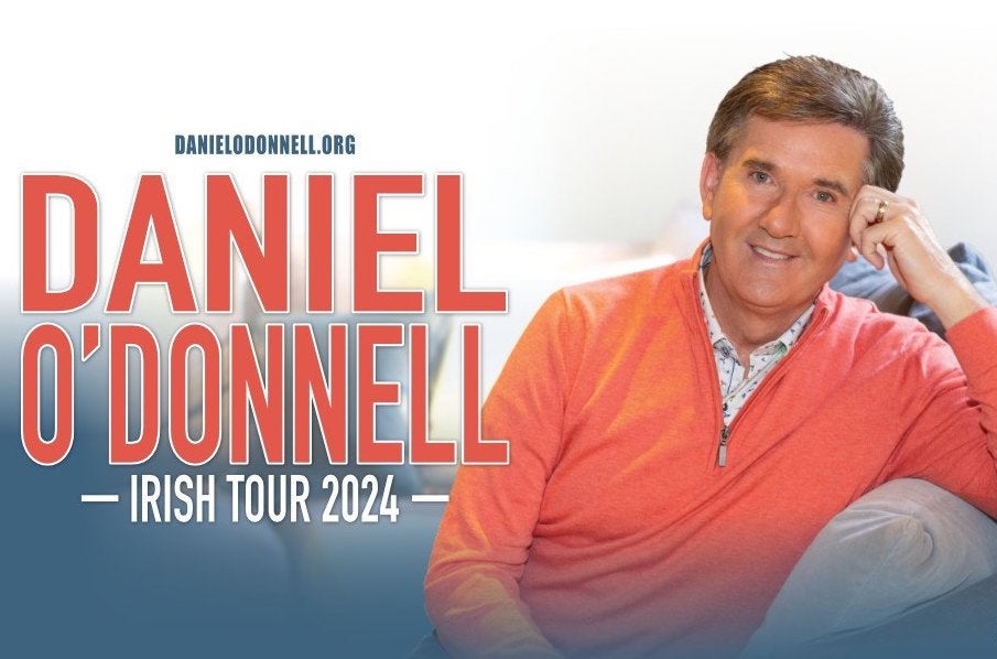 Daniel O'Donnell in concert. A gently smiling man in an orange jumper is resting his left arm on the arm of a sofa.
