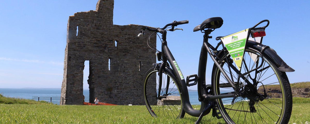 A lone bicycle propped up with the ruins of a small castle a short distance away with the coast in the background