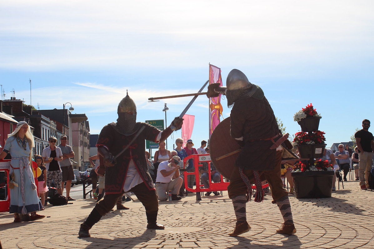 2 men dressed in viking type clothing in a mock battle with swords on a paved area with people watching in the background and blue sky.