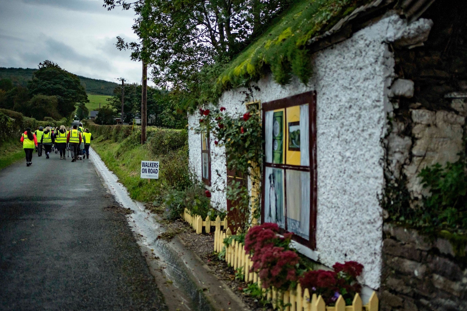 Looking down a country lane with group in hi-vis jackets walking away and old cottage in the foreground.