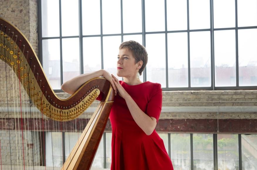 A woman in a red dress is leaning on a large harp looking away to her right.
