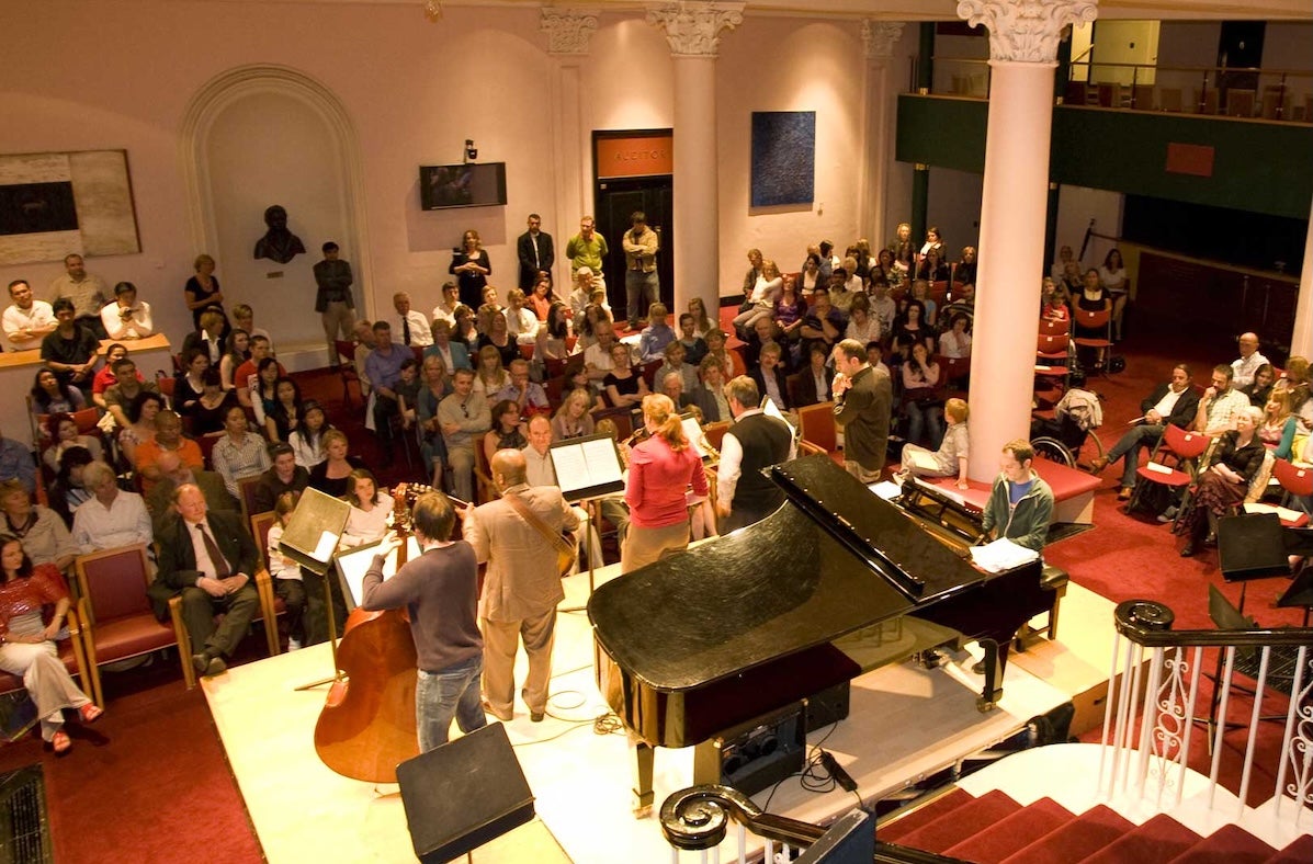 Viewed from behind and above, rear view of small stage with musicians playing and seated audience in a hallway with stairs