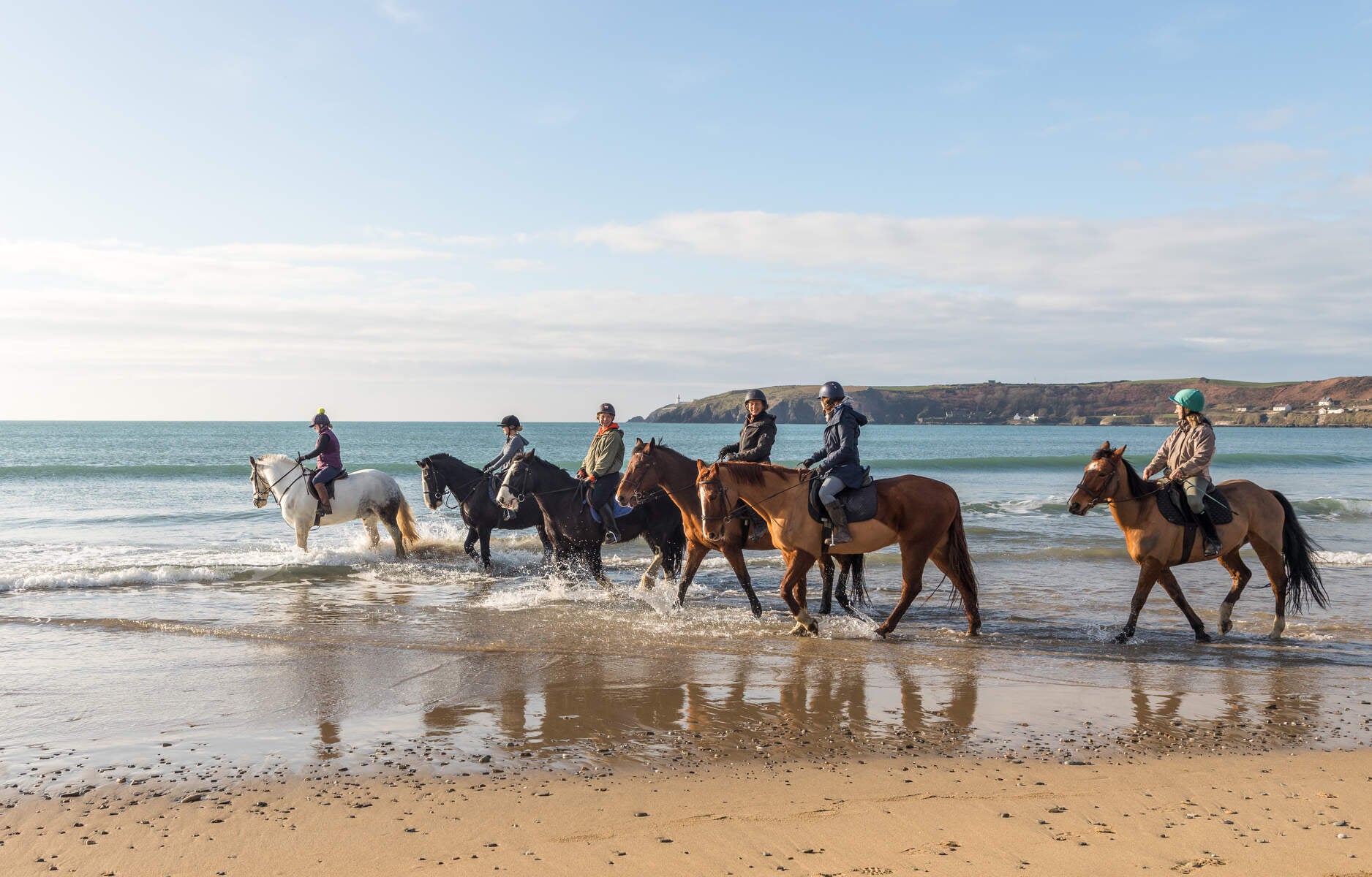 Horses and riders in the water under a blue sky with sand to the forefront