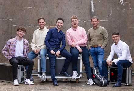 Line of 6 smiling men in different coloured shirts, seated casually on equipment boxes.