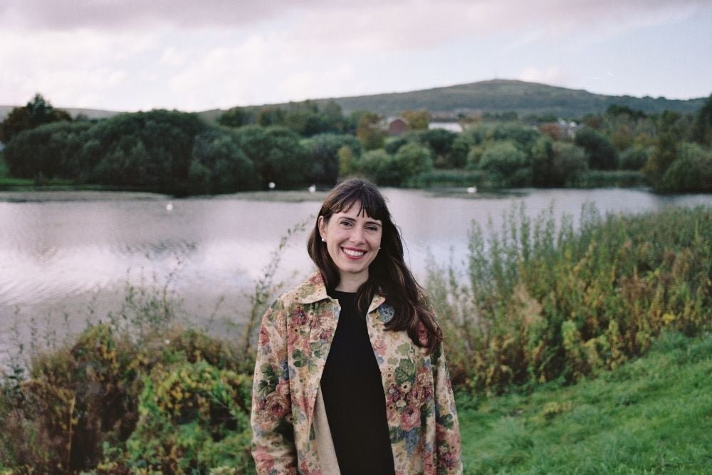 Photo Mitch Conlon. A smiling woman outdoors with a lake and bushes behind her.