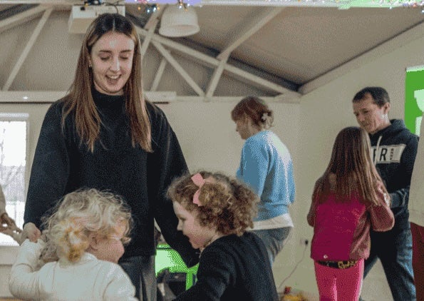 pop-up Gaeltacht at Ardgillan Castle - a smiling woman looking down on 2 young children all dancing in a circle with others in the background.
