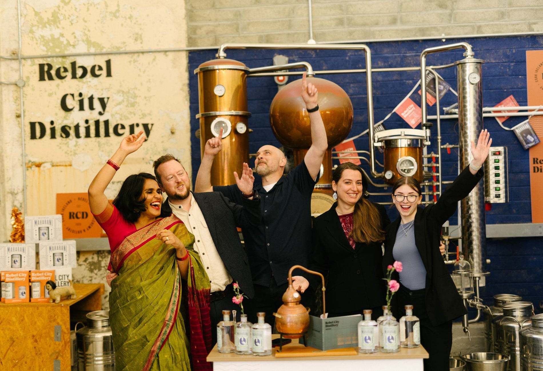 Five people with raised arms standing in a distillery and a counter with bottles on it in front