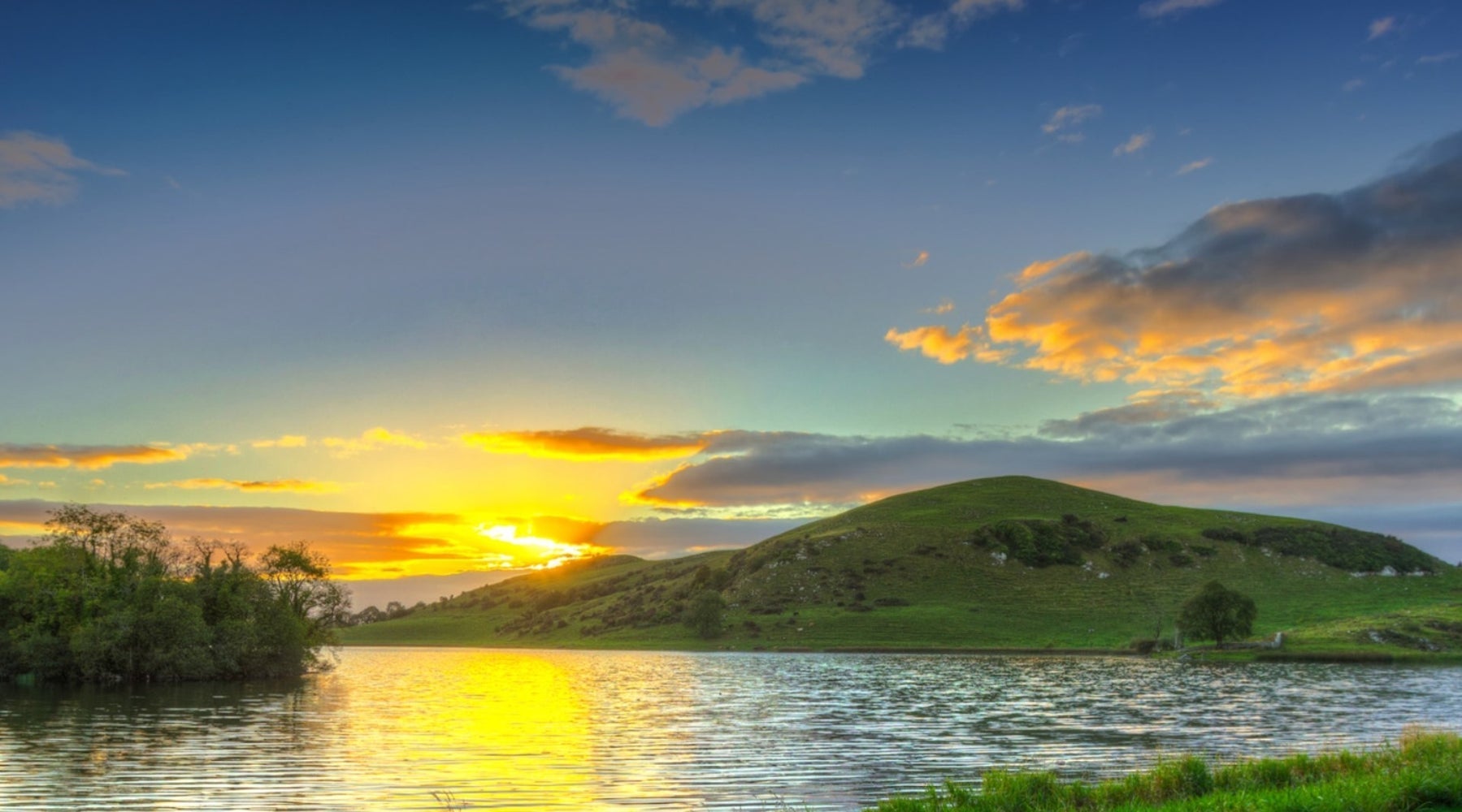 Sun setting behind green hills beside a lake