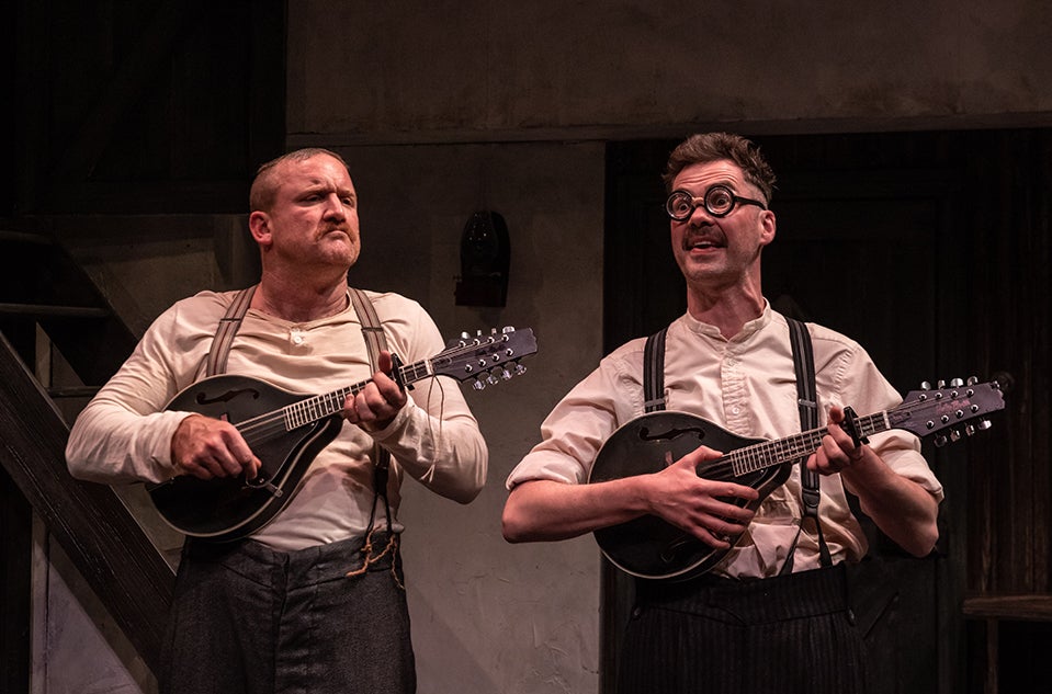 DRUID - Three Short Comedies at Watergate Theatre. 2 men in old style collarless shirts are playing banjos with animated faces on a stage.