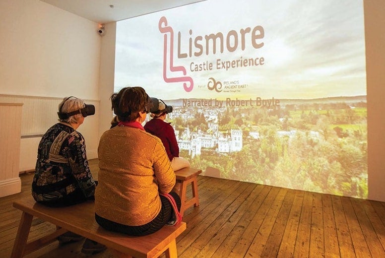Rear view of 3 people wearing VR sets sitting on small wooden benches facing a large image projected onto a blank wall.