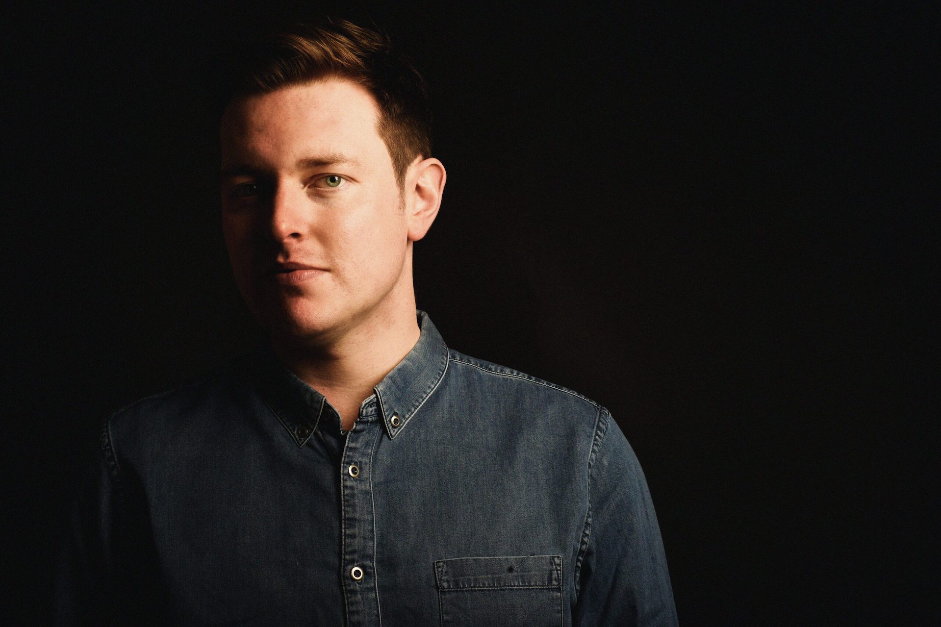 A man in dark denim shirt is looking at the camera in partial shadow against black background