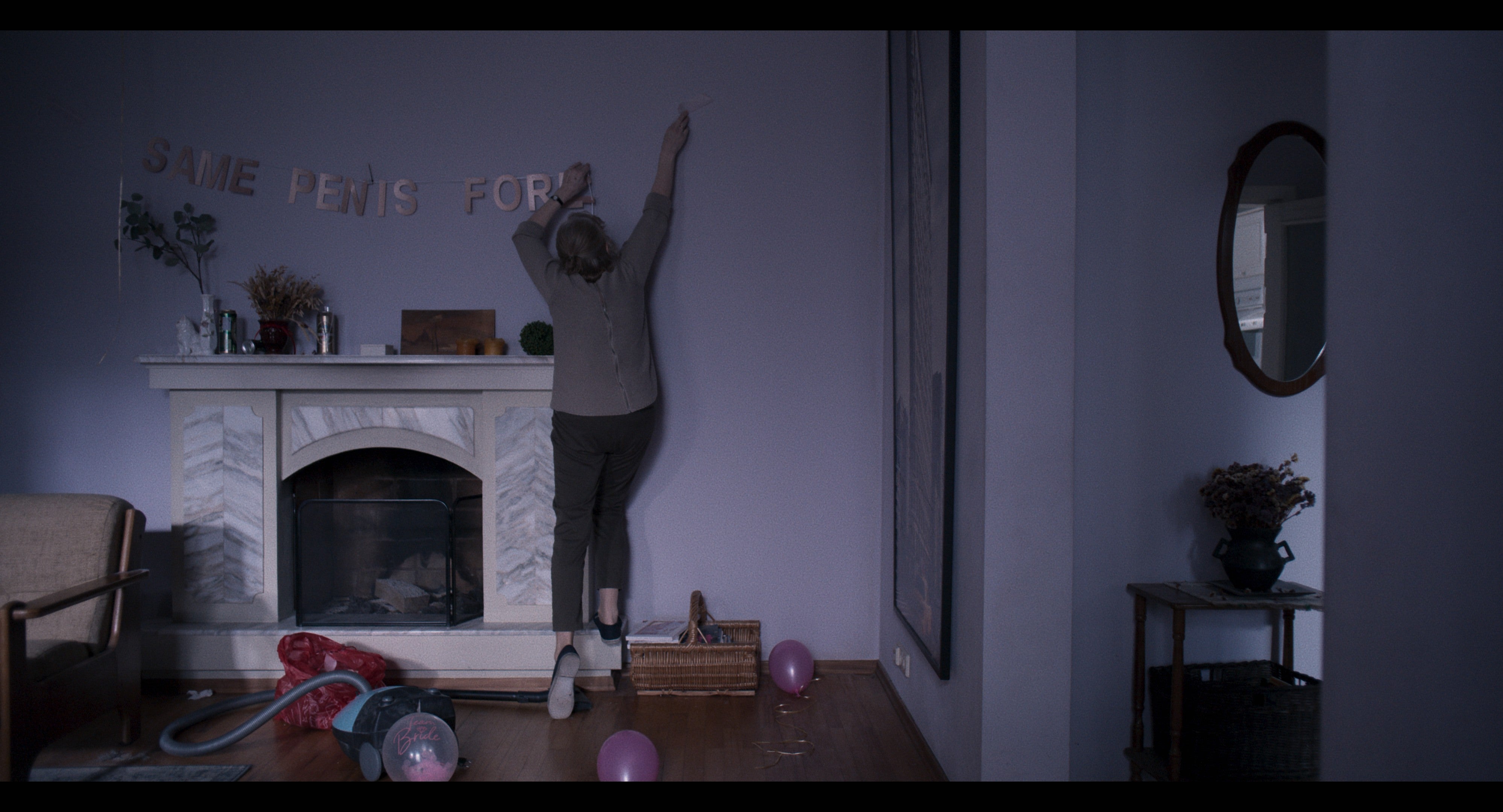 A woman hanging up letters that spell "Same Penis For" above a fireplace. The third word is apparently incomplete.