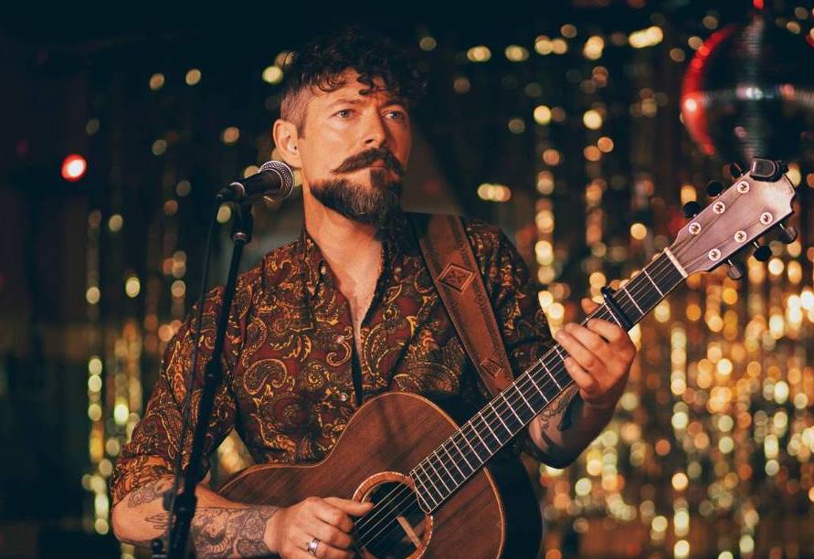 Niall McCabe to play Gleneagle Arena- a man playing a guitar standing at a mic on on stand with dark and gold blurry background.
