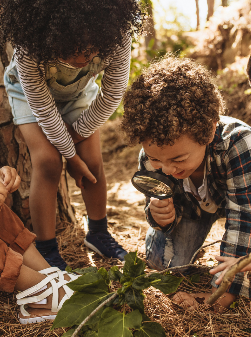 2 young children, one looking through a magnifying glasses are looking down at leafs and brown matter on the ground.