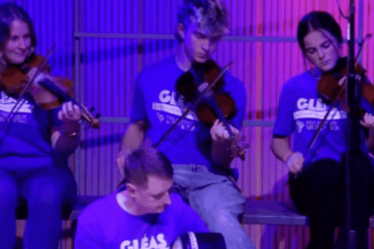 A group of 4 musicians playing fiddles in blue T-shirts lit by purple light