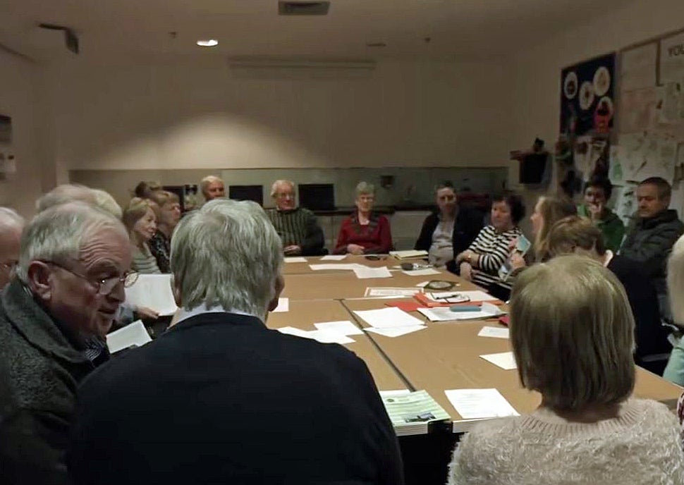 People seated around a large table with papers on listening to a woman talking.