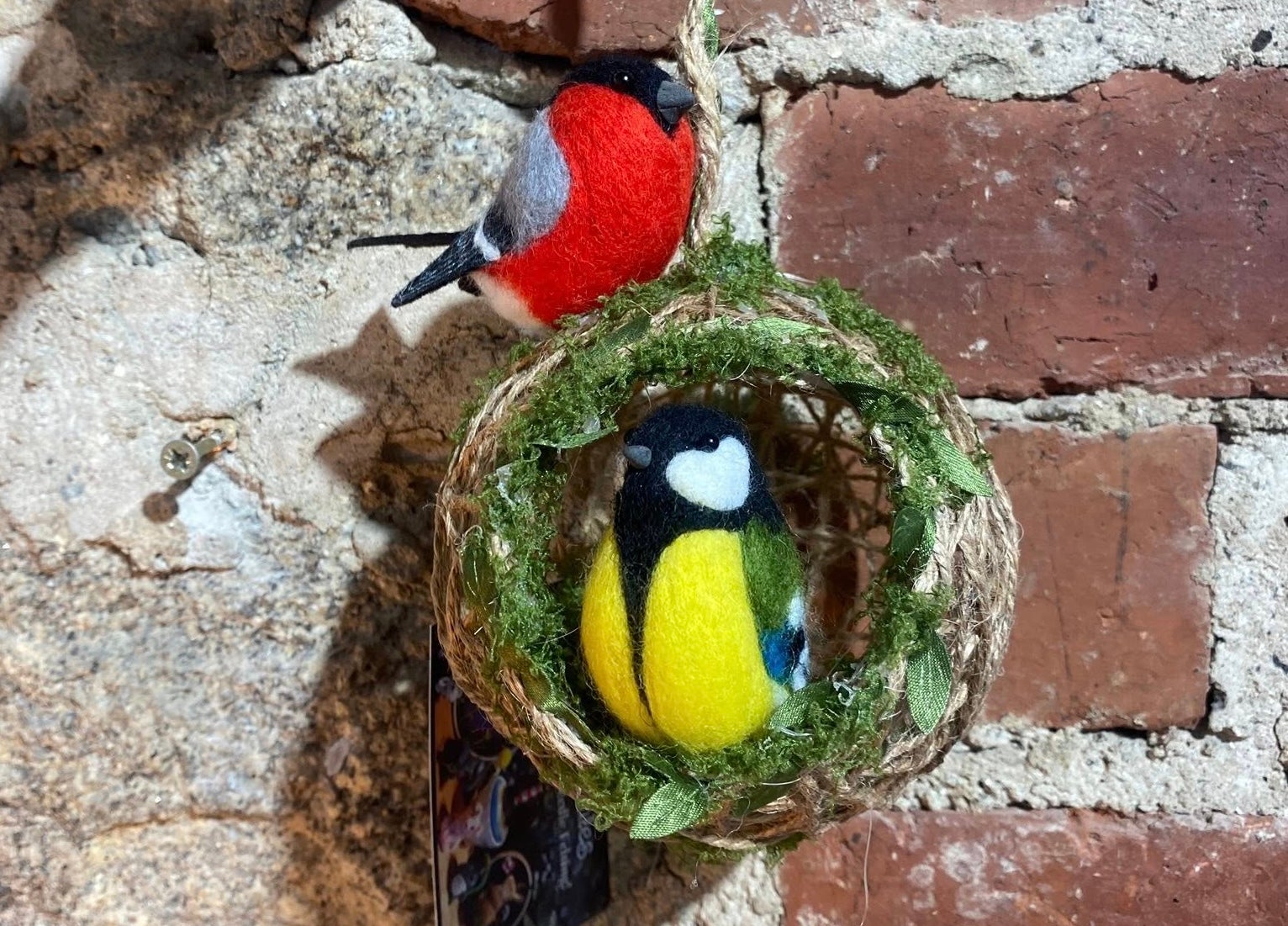 A felt brightly coloured bird in a small round woven half open ball with another felt brightly coloured bird on the top.