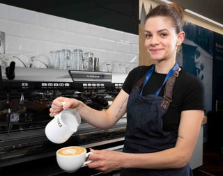 Person holding a milk frother and a cup of coffee while standing in front of coffee machine