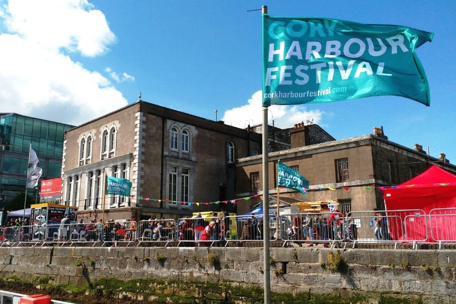 Cork Harbour Festival 2025, blue flag flying on a quay with stone buildings and people.