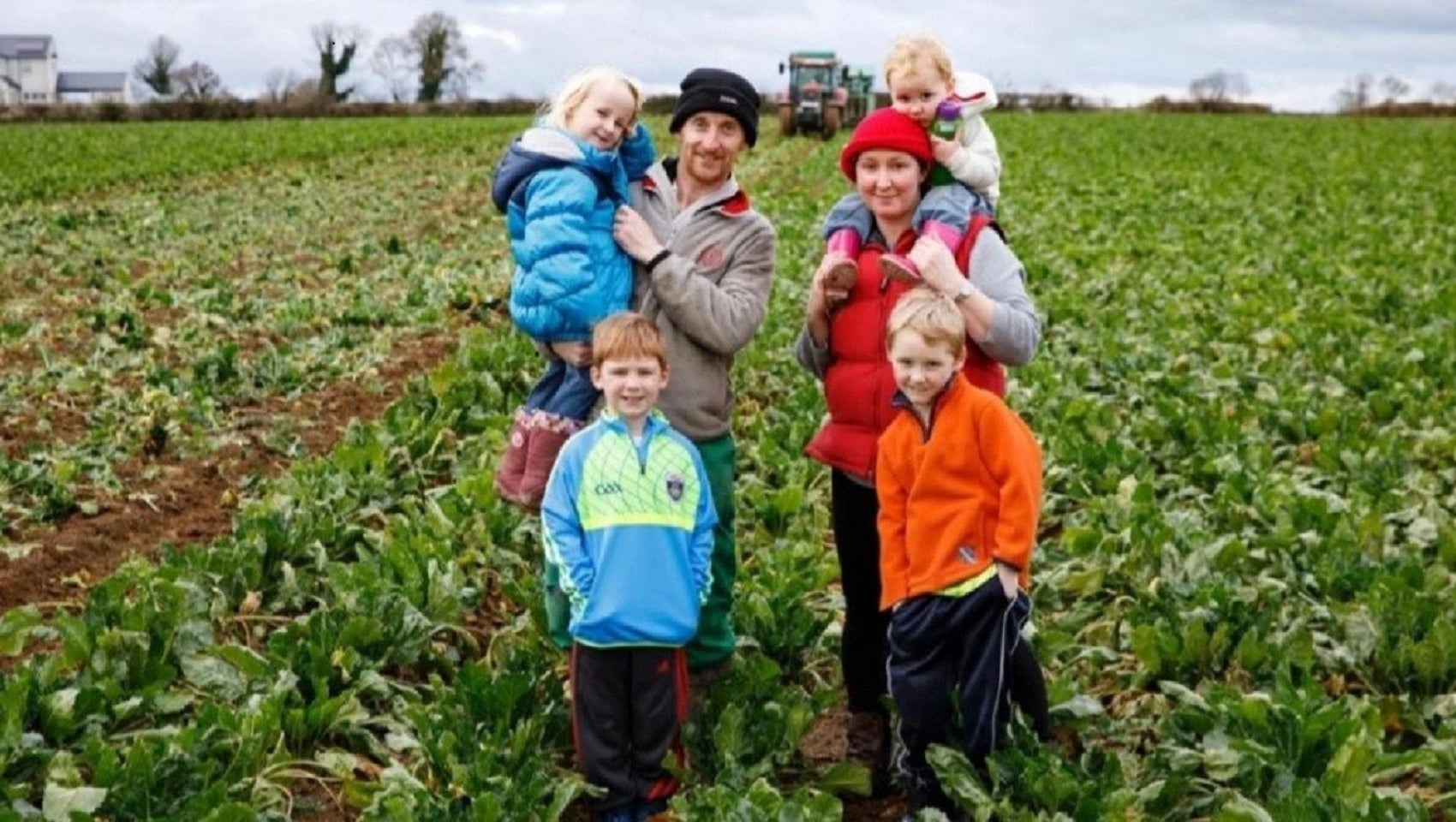 Group of adults and kids standing in a field with a tractor in the background