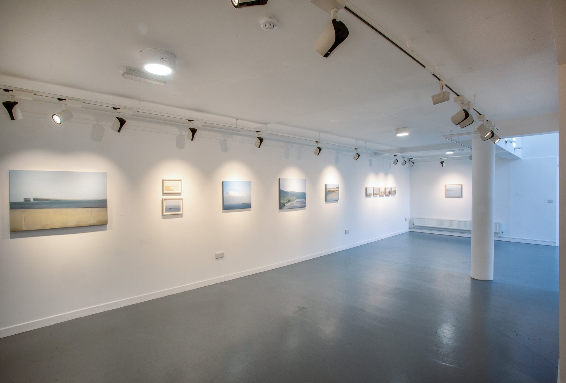 Gallery with dark wooden floors a white column and white walls with art displayed on them