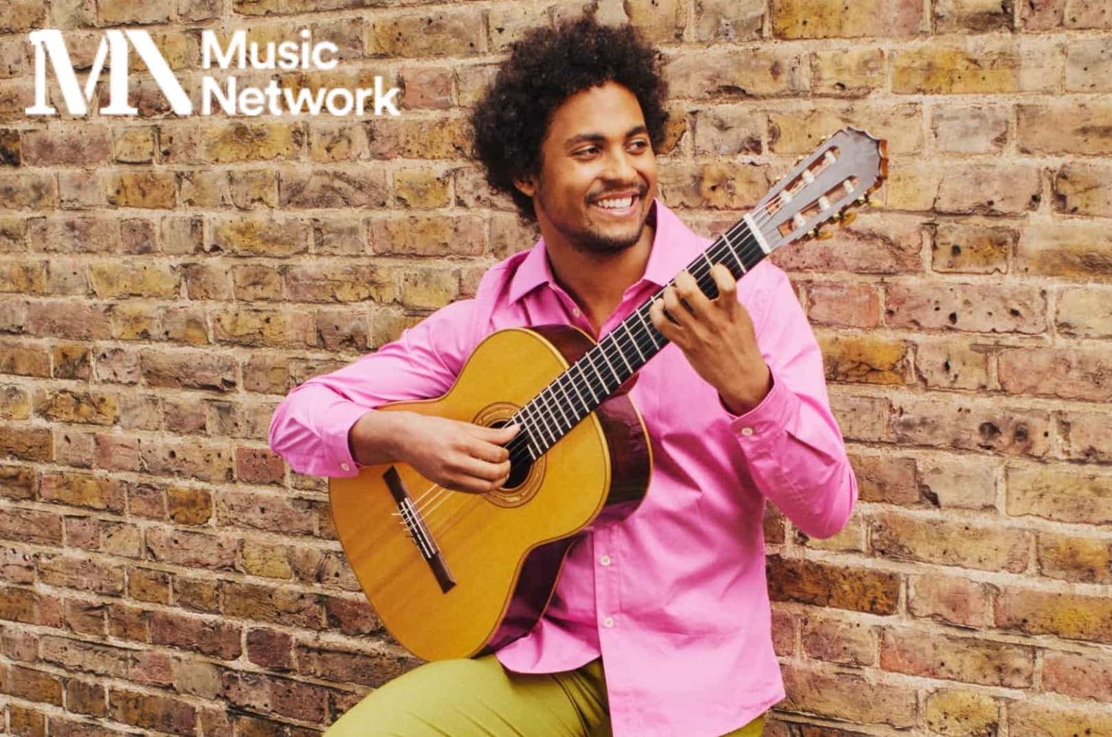 A smiling man in pink shirt is playing a guitar, leaning against a yellow brick wall.