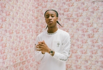 A man in white top is standing with hands held together in front of him and background of pink patterned walls.