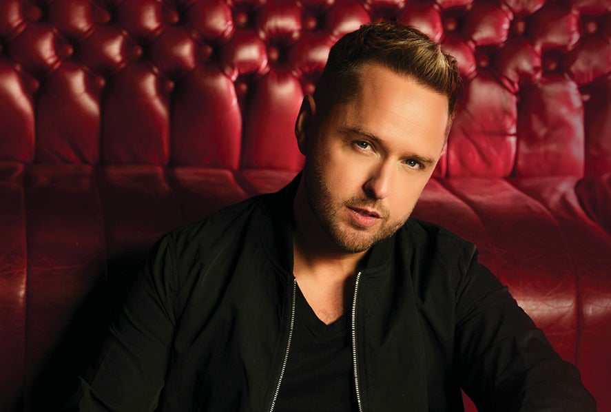 A man in black top and jacket is looking serious, against background of a burgundy leather bench seat