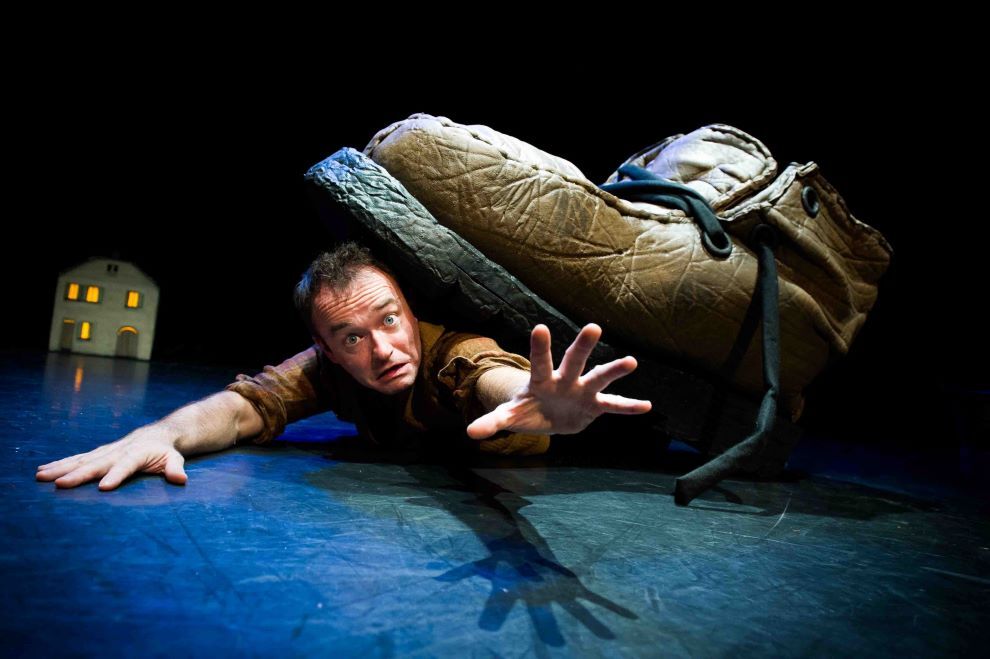 An anxious looking man is sprawled out on a stage floor reaching out with a very large boot on his back.